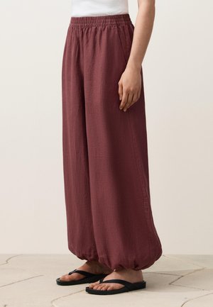 Luźne bordowe spodnie z elastycznym pasem i marszczonymi mankietami, noszone z czarnymi sandałami i białym topem, pokazane od talii w dół.