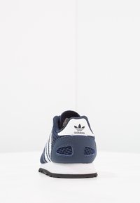 Marinblå sportskor med mesh-överkärl, vita ränder, vadderad krage och svart gummisula. Har en logotyp på hälen.