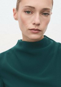 Primer plano de una persona que lleva una prenda de textura suave de color verde oscuro con un cuello alto, hecha de un tejido suave y fluido. Fondo neutro.