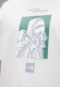 Λευκό βαμβακερό t-shirt με πράσινο και γκρι γραφικό που απεικονίζει μια φιγούρα με κουκούλα. Περιλαμβάνει το λογότυπο "THE NORTH FACE" στην κάτω γωνία.