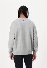 Sweatshirt cinza com um ajuste solto, punhos e bainha canelados, apresentando um pequeno logótipo azul da Nike nas costas. Textura suave, design clássico.