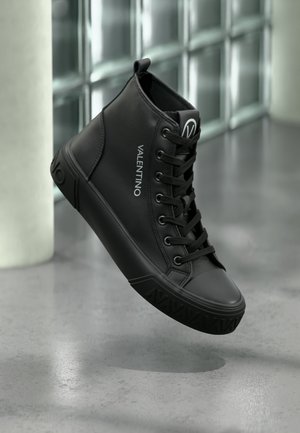 Zapatilla negra de caña alta con cordones, logo "VALENTINO" en el lateral, detalle de suela en relieve, presentada sobre un fondo moderno de bloques de vidrio.