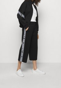 Czarna kurtka bomberka z detalem logo, biała bluzka, czarne spodnie 3/4 z bocznym paskiem z logo oraz białe trampki. Gładka tekstura tkaniny.