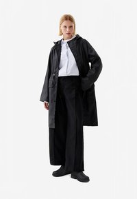 Μαύρο αδιάβροχο παλτό με κουκούλα, φορεμένο πάνω από μια λευκή ριγέ πουκάμισο, συνδυασμένο με φαρδιά μαύρη παντελόνια και μαύρα παπούτσια slip-on.