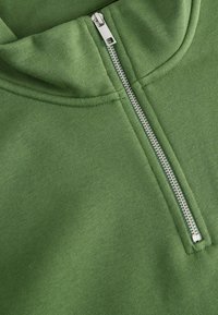 Green sweatshirt with a high collar and a silver zipper. Smooth texture with stitching details along the collar and sides.