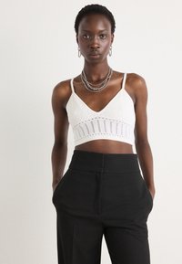 Weißes Spitzen-Bralette mit V-Ausschnitt und verstellbaren Trägern, das mit einem strukturierten Muster und Ausschnitten versehen ist, kombiniert mit hochgeschnittenen schwarzen Hosen.