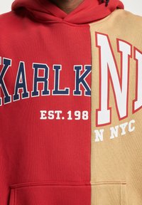 Červená a béžová mikina s rozdeleným dizajnom. Obsahuje nápis "KARLK" v námorníckej modrej farbe a "NYC" v bielej. Mäkká textúra a klokania kapsa.