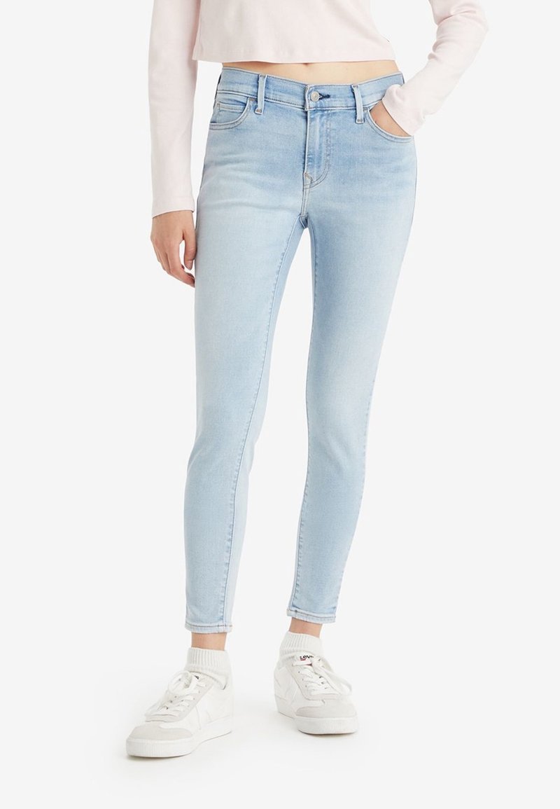 Woman wearing light blue skinny jeans, white socks, and white sneakers, with one hand in pocket against plain background.