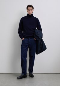 Pull à col roulé marine, jean en denim foncé et bottes en cuir noir. Le modèle tient une veste foncée, debout devant un fond neutre.