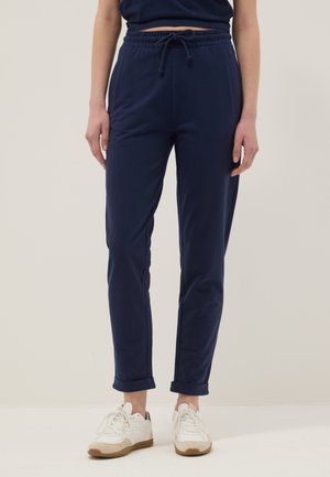 Frau trägt marineblaue Hosen mit Kordelzug, an den Knöcheln hochgekrempelt, und weiße Sneakers, steht vor einem einfarbigen hellen Hintergrund.