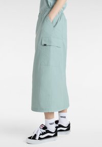 Light green cargo skirt with side pockets and a relaxed fit, made of textured fabric. Paired with black and white sneakers and socks.