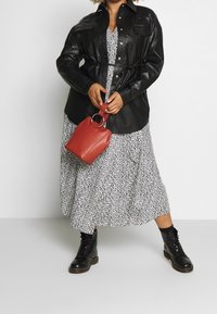 Černá kožená košile-jacket s kapsami, s páskem v oblasti pasu. Pod tím je bílá šaty s černými tečkami. Černé šněrovací boty dotvářejí celkový vzhled.