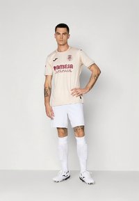 Beež jalgpallivorm punase logoga ja tekstiga, lühikeste varrukatega ning tekstuursete triipudega; kombineeritud valgete pükste ja sokkidega ning mustade ketsidega.