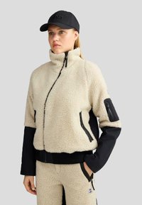Giacca in fleece color crema con maniche nere, colletto alto e tasche con zip. Caratterizzata da una texture morbida e un design sportivo.