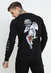 Sudadera negra de manga larga con un gráfico detallado de un querubín sosteniendo rosas en la parte posterior. Texto blanco a lo largo de la manga.
