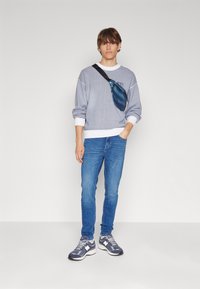 Sweatshirt gris en tricot avec des poignets blancs, jean bleu et baskets grises. Un sac bandoulière teal repose sur l'épaule. Tenue simple et décontractée.