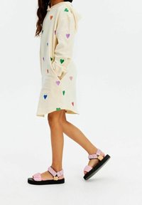 Donna che cammina in un vestito a hoodie di colore crema a lunghezza ginocchio con motivi a cuori colorati, indossando sandali rosa con tracolla testurizzata e suole nere.