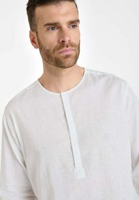 Man met kort bruin haar en baard, gekleed in een wit, knoopshirt van linnen, kijkt lichtjes opzij tegen een effen achtergrond.