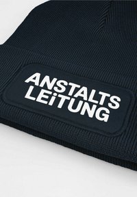 Navy knitted beanie with a ribbed texture and a rectangular patch featuring the text "ANSTALTSLEITUNG" in bold white letters.