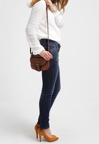 Brun lädercrossbody-väska med rundad form, prydd med en metallknäppning och en lång rem; bärs med navy skinny jeans och orange klackar.