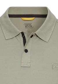Lys grøn polo skjorte med ribbet krave, to sorte knapper og en kontrasterende gul detalje på indersiden af kraven. Har logoet "camel active."
