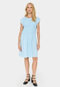 Ljusblå klänning gjord av mjukt tyg, med en smockad bodice, korta ärmar och enfladdrig kjol. Matchas med svarta klackskor med blockklack.
