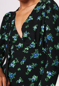 Robe noire à motifs floraux avec des roses bleues et des feuilles vertes, présentant un décolleté en V profond et des manches bouffantes. Le tissu semble doux et texturé.