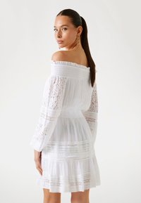 Abito bianco off-shoulder con dettagli in pizzo, maniche lunghe a sbuffo, vita arricciata e orlo a balze stratificate. Tessuto liscio e leggero.
