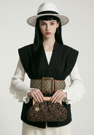Jeune femme portant un chapeau blanc, un gilet matelassé noir, une large ceinture à motif, tenant une pochette marron à motif géométrique.