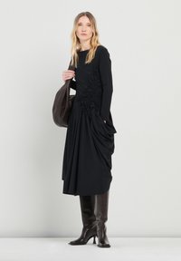 Vestido negro de manga larga con detalles fruncidos en el costado, una silueta fluida, combinado con botas de tacón marrones hasta la rodilla y un bolso marrón.