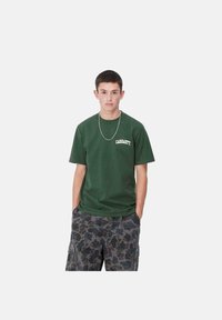 Camiseta de algodón verde oscuro con cuello redondo y logo blanco de "Carhartt" en el lado izquierdo del pecho, combinada con pantalones cortos cargo de patrón de camuflaje.