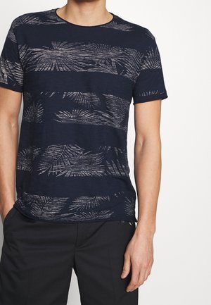 Man in een marineblauw hemd met korte mouwen en lichte grijze tropische bladerenpatronen, met zwarte broek, staand met handen in zijn zakken.