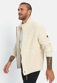 Knit cardigan in cream, featuring a structured design, high collar, front buttons, and side pockets. Textured fabric with ribbed cuffs.