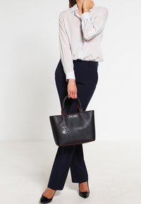 Kvinna i vit skjorta och marinblå byxor, som håller en svart designerväska med metall-logotyp, stående mot en neutral bakgrund.