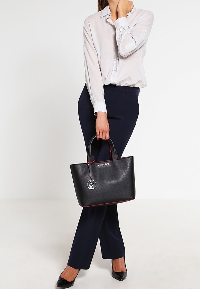 Kvinna i vit skjorta och marinblå byxor, som håller en svart designerväska med metall-logotyp, stående mot en neutral bakgrund.