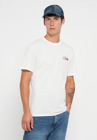 Hvit bomull t-skjorte med rund hals og korte ermer, med rød brodert tekst på brystet. Kombinert med blå denim jeans.