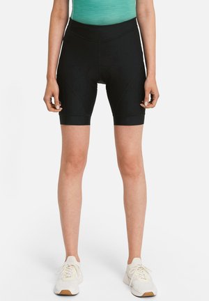 Woman wearing black padded cycling shorts, teal top, and white sneakers, standing against a plain white background.