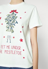 Vit bomull T-shirt med en mistelgröna design och ett rött band, omgiven av gröna och röda stjärnor. Texten lyder: "MÖT MIG UNDER MISTELN."