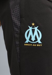 Material sport negru cu logo albastru deschis Olympique de Marseille, un stea aurie deasupra și textul „DROIT AU BUT” dedesubt.