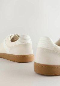 Hvide sportssko med en glat overflade, tan gummisål og polstret hældetalje. Har syede detaljer og teksturerede overlays.