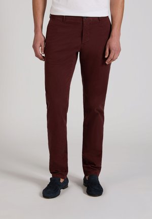 Man met slim fit donkerrode broek en marineblauwe suède loafers, staand tegen een effen lichtgrijze achtergrond.