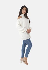 Femme portant un pull en tricot blanc à épaules dénudées, un jean skinny bleu et des sandales à talons transparents, se tenant les mains jointes.