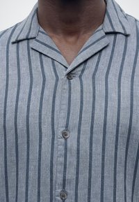 Detail tmavé osoby v šedém tričku s vertikálními tmavě modrými pruhy a třemi viditelnými knoflíky.