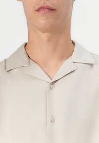 Short-sleeve, beige shirt with a smooth texture, a classic spread collar, and two buttons at the placket. No patterns or accents visible.