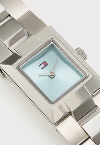 Reloj de metal plateado con una caja rectangular, esfera azul clara, manecillas de horas y minutos plateadas, y un pequeño detalle del logo de una bandera.