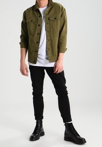 Veste en denim vert olive avec deux poches avant, t-shirt blanc en dessous, pantalon noir ajusté, et bottes noires à lacets.
