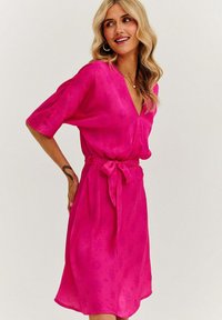 Fuksiová šaty s výstrihom do V, krátkymi rukávmi a opaskom na zaväzovanie. Ľahký materiál s jemnými kvetinovými vzormi a splývavým lemom.