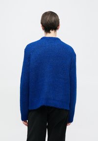 Kék kötött pulóver laza szabással, bordázott textúrával és nyers szegéllyel. Kendőgallérral és hosszú ujjakkal.