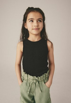 Schwarzes, strukturiertes Tanktop kombiniert mit hellgrünen, rüschelangen Shorts mit einem elastischen Bund und seitlichen Taschen. Das Kind steht mit den Händen in den Taschen.