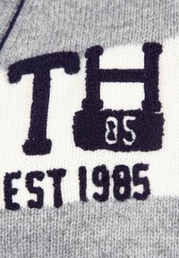 Wykonana na drutach tekstylia w odcieniach szaro-białych, z haftowanym, granatowym tekstem: "TH", "85" oraz "EST 1985". Tekst ma lekko wypukłą teksturę.